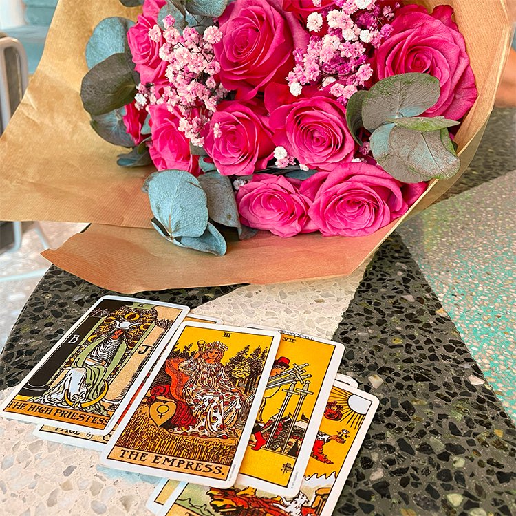 A bouquet of pink roses with greenery and babys breath rests on a speckled tabletop beside four tarot cards The High Priestess