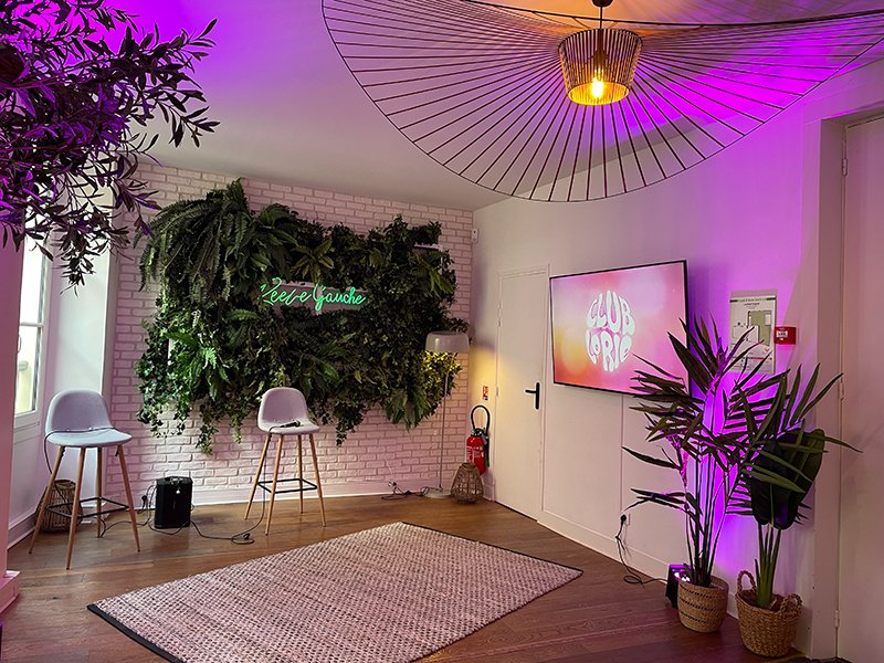 A modern room with a green plant wall, two chairs, a rug, and a TV displaying “CLUB LORIE.” Pink and purple lighting adds ambi