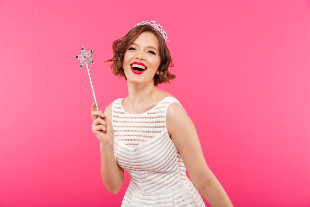 Portrait of a laughing girl wearing crown and holding magic wand while looking at camera isolated over pink background