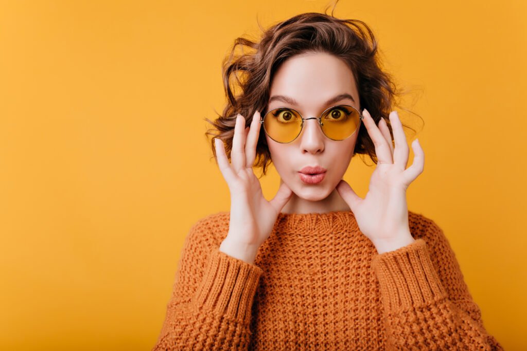 Close-up portrait of white funny woman in vintage yellow glasses. Indoor photo of blissful curly caucasian girl in soft sweater isolated on bright background..