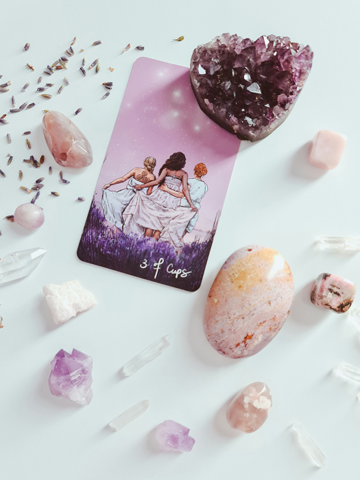 A tarot card labeled 3 of Cups with three women in white dresses, surrounded by crystals, stones, an amethyst cluster, and s